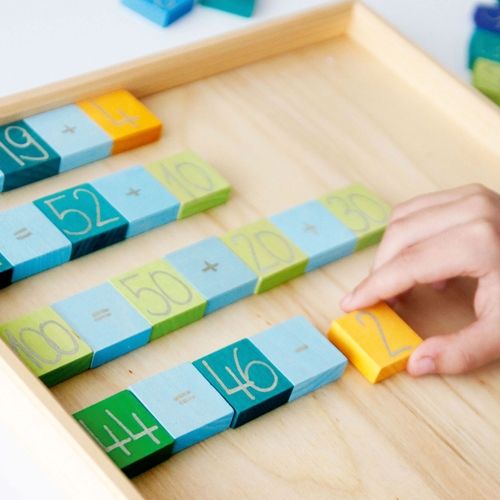 Een kinderhand plaatst een geel cijferblokje in een houten bak met grimm's rekenblokjes.