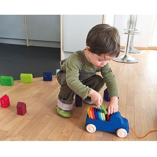 Een kind speelt met de Grimm's houten trekfiguur vrachtwagen en regenboogkleurige blokken op een houten vloer.