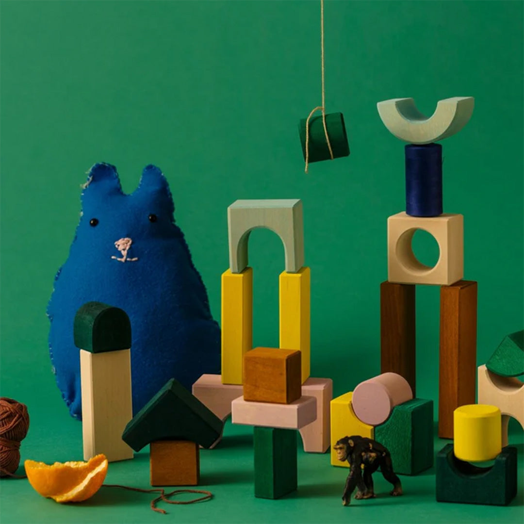 Een creatieve opstelling van Grimm's veelkleurige houten bouwblokken met een blauwe pluche kat en een speelgoedaapje op een groene achtergrond.