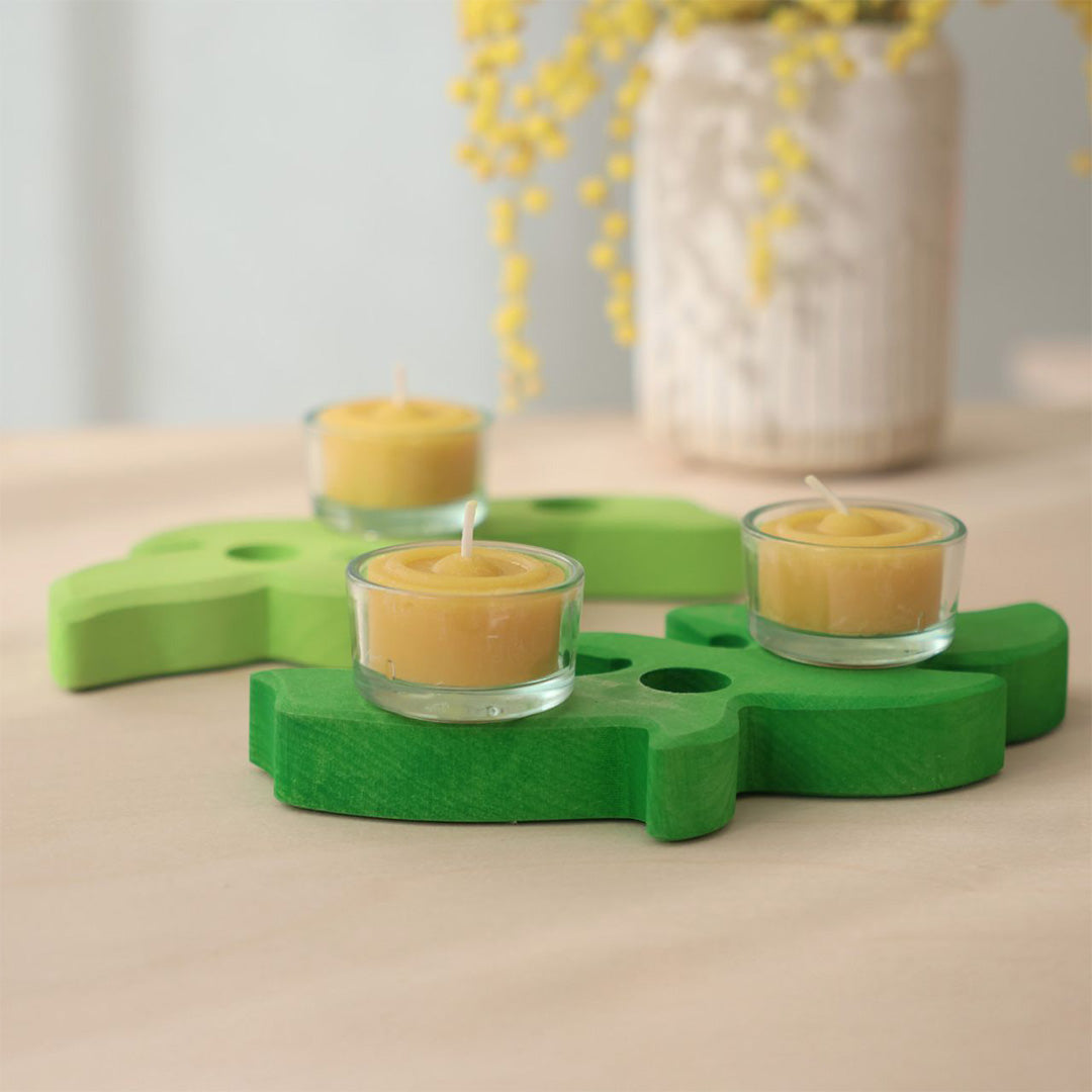 Twee Grimm's glazen houders met gele waxinelichtjes op groene houten decoratiestukken.