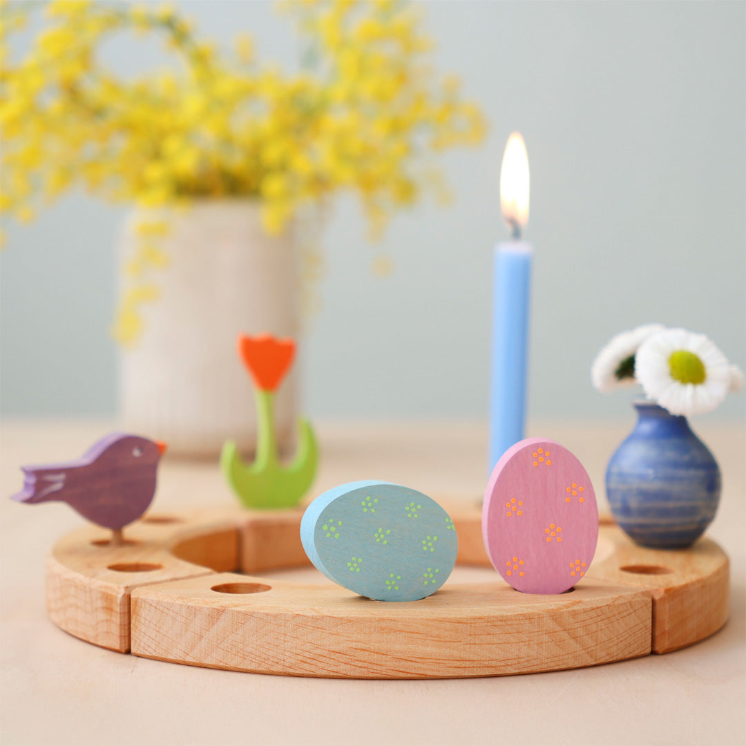 Twee Grimm's houten paaseieren decoratie figuren in een houten ring met andere paasdecoraties en een brandende kaars.