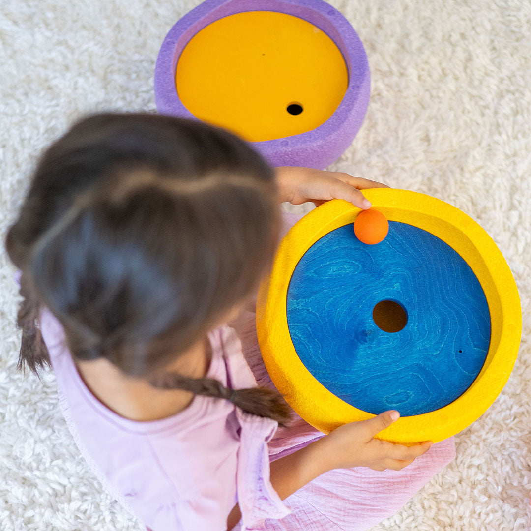 Een kind speelt met een gele grimm's x stapelstein rolling disc met een blauwe houten schijf en een oranje bal.