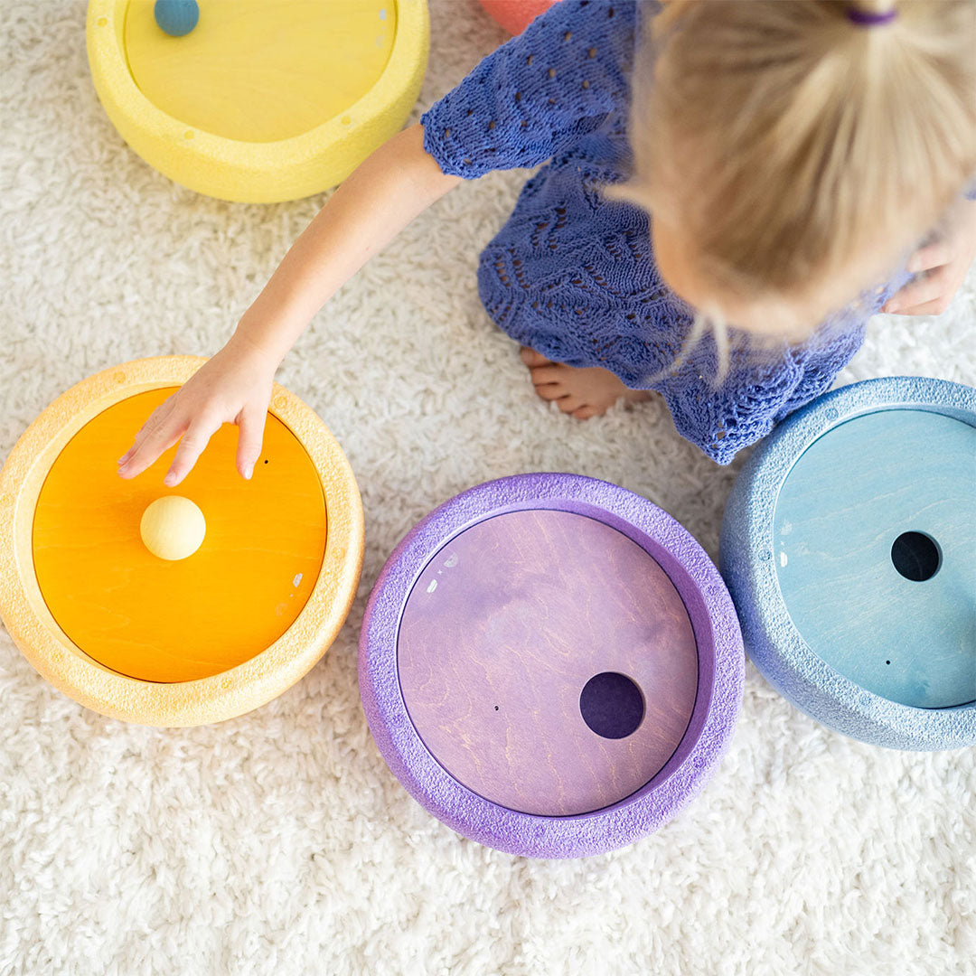 Bovenaanzicht van een kind dat op een wit vloerkleed speelt met grimm's rolling discs in pastelkleuren, waarbij een hand een gele disc met een balletje aanraakt.