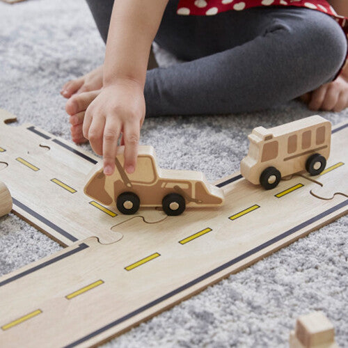 Een kind speelt met een houten bulldozer en bus op een houten autoweg met een kruispunt.
