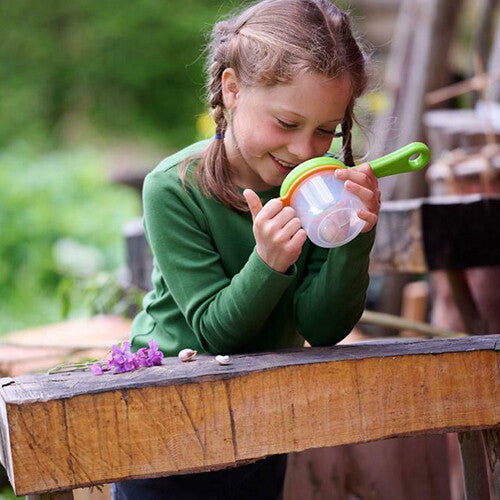Een lachend kind bekijkt kleine voorwerpen in de Haba bekerloep, buiten op een houten balk.