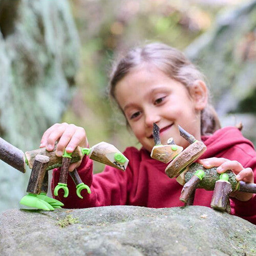 Een lachend meisje speelt buiten met twee zelfgemaakte dinosauriërs van takken en groene connectors.