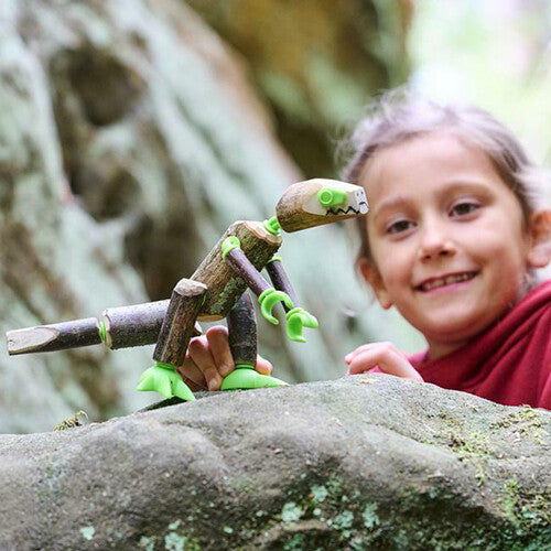 Een lachend meisje kijkt naar een zelfgemaakte dinosaurus van takken en groene connectors op een rots.