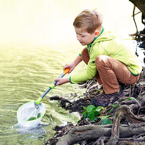 Een jongen in een groene trui vangt waterdiertjes met het Haba schepnetje in een beekje.