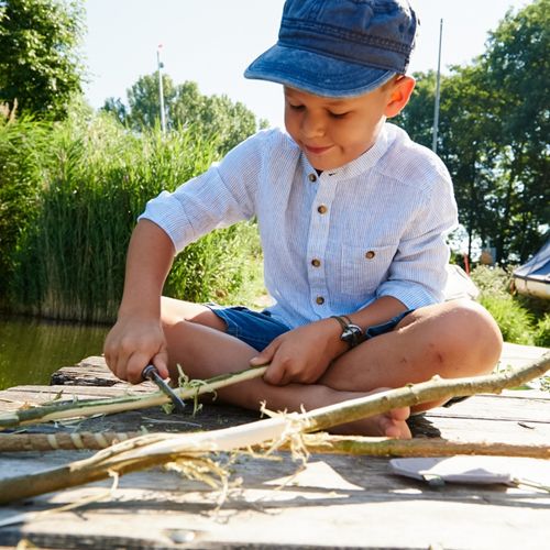 Een jongen pelt met een Haba Terra Kids zakmes de bast van een tak op een houten vlonder aan het water.