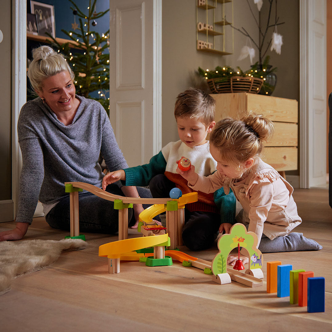 Een vrouw en twee kinderen spelen samen met de HABA Kullerbü knikkerbaan op een houten vloer.