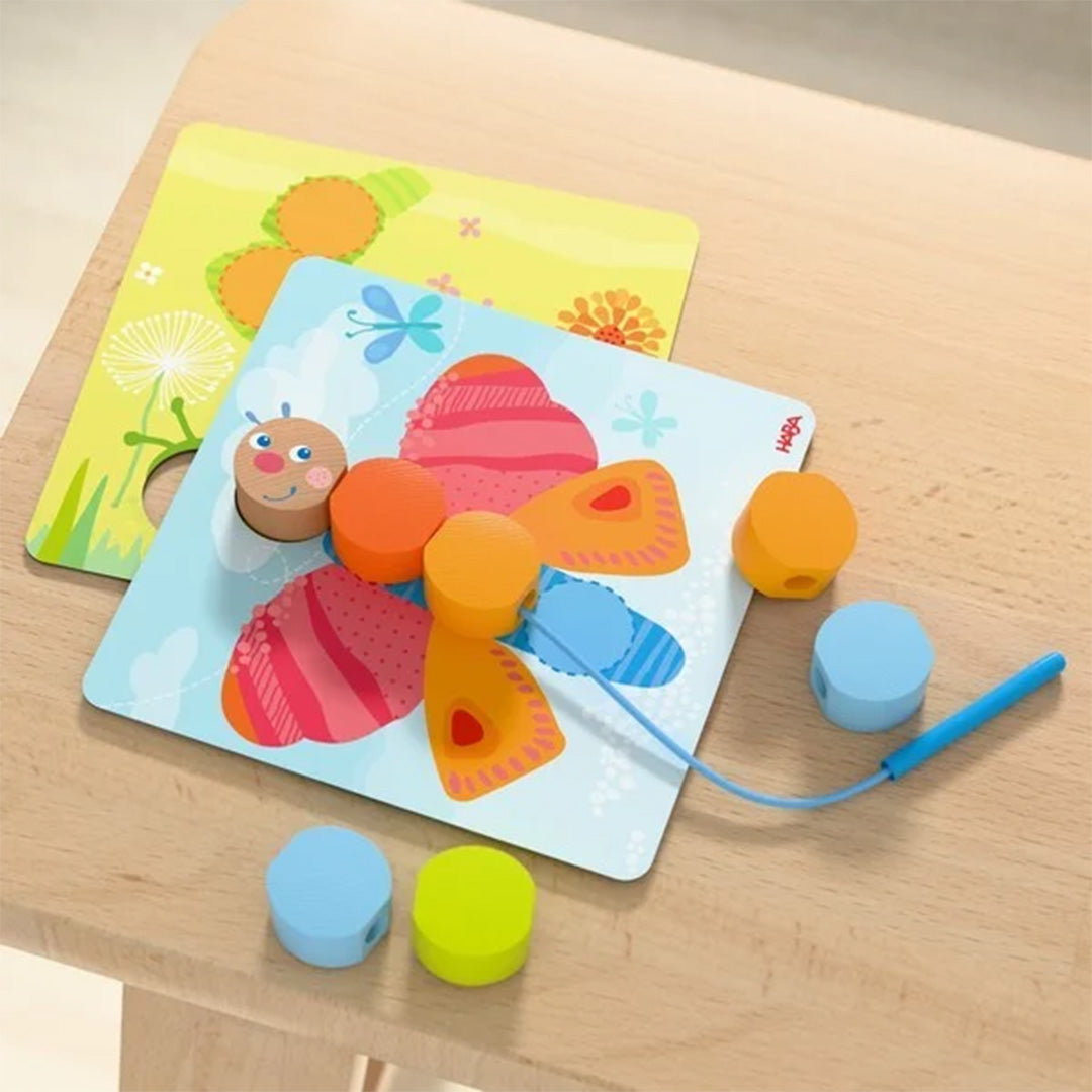 HABA rijgspel met houten rups en vlinder op een blauwe kaart, met losse rijgkralen en een rijgnaald.