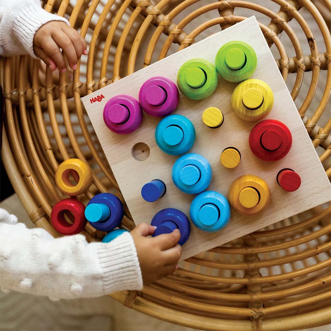 Twee kinderhanden sorteren veelkleurige houten ringen en staafjes van het Haba sorteerspel op een rieten tafel.
