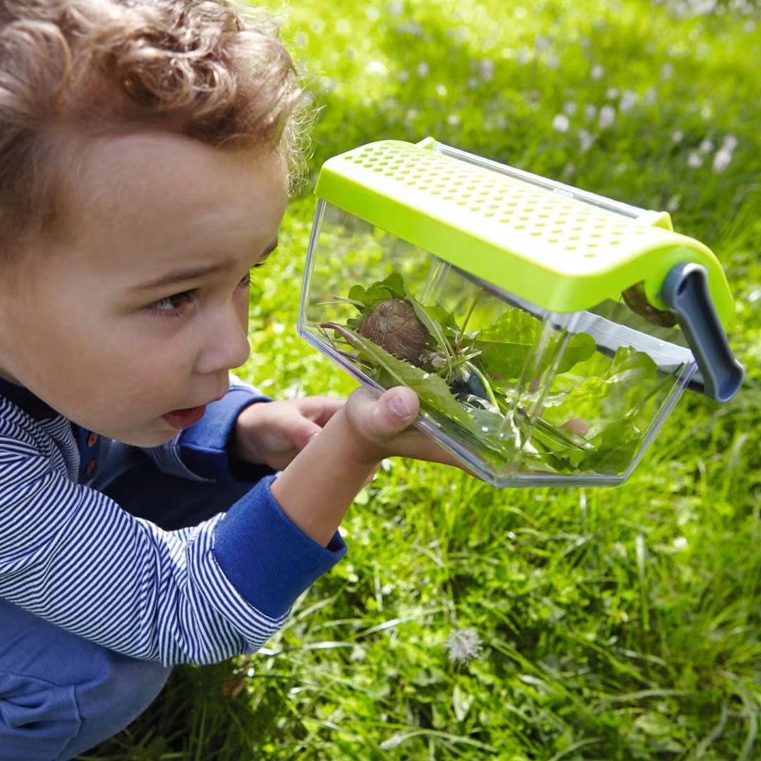 Een kind kijkt geconcentreerd naar de Haba insectendoos met een slak en bladeren erin, zittend in het gras.
