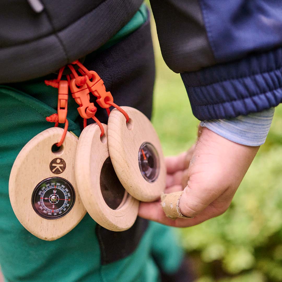 Een kinderhand houdt de Haba Terra Kids ontdekkingsset met kompas, thermometer en loep vast, bevestigd aan een groene broek.