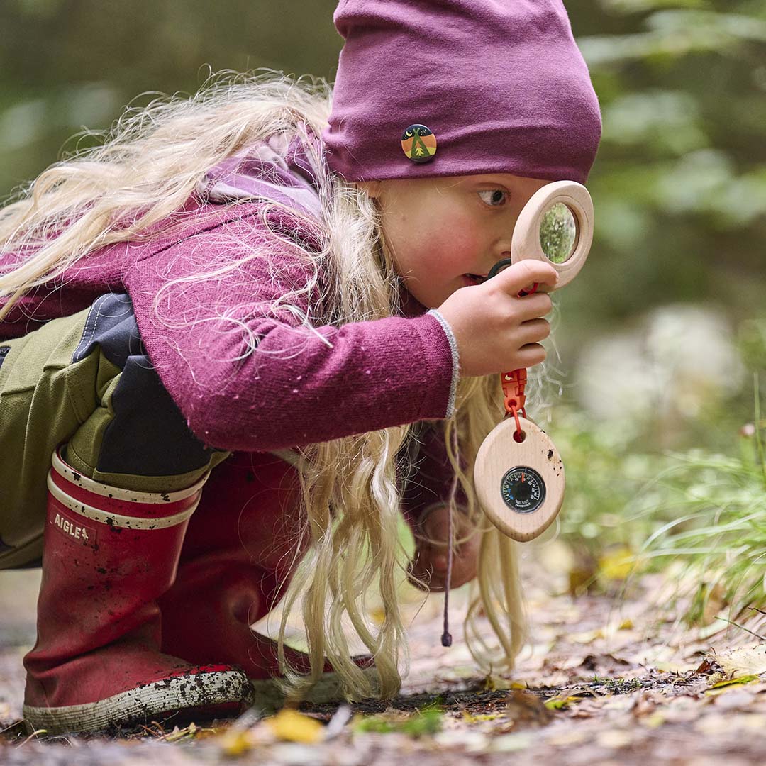 Een meisje in het bos kijkt door de loep van de Haba Terra Kids ontdekkingsset naar de grond.