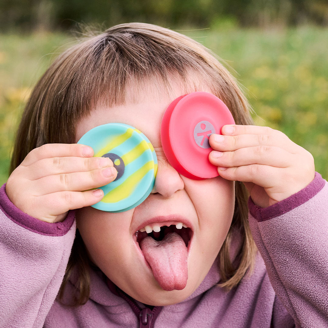 Een lachend kind met een turquoise-gele en een rode mini frisbee voor haar ogen.