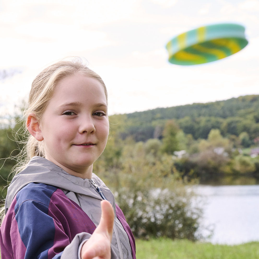 Een lachend meisje speelt buiten met een Haba Terra Kids gestreepte mini frisbee die in de lucht zweeft.