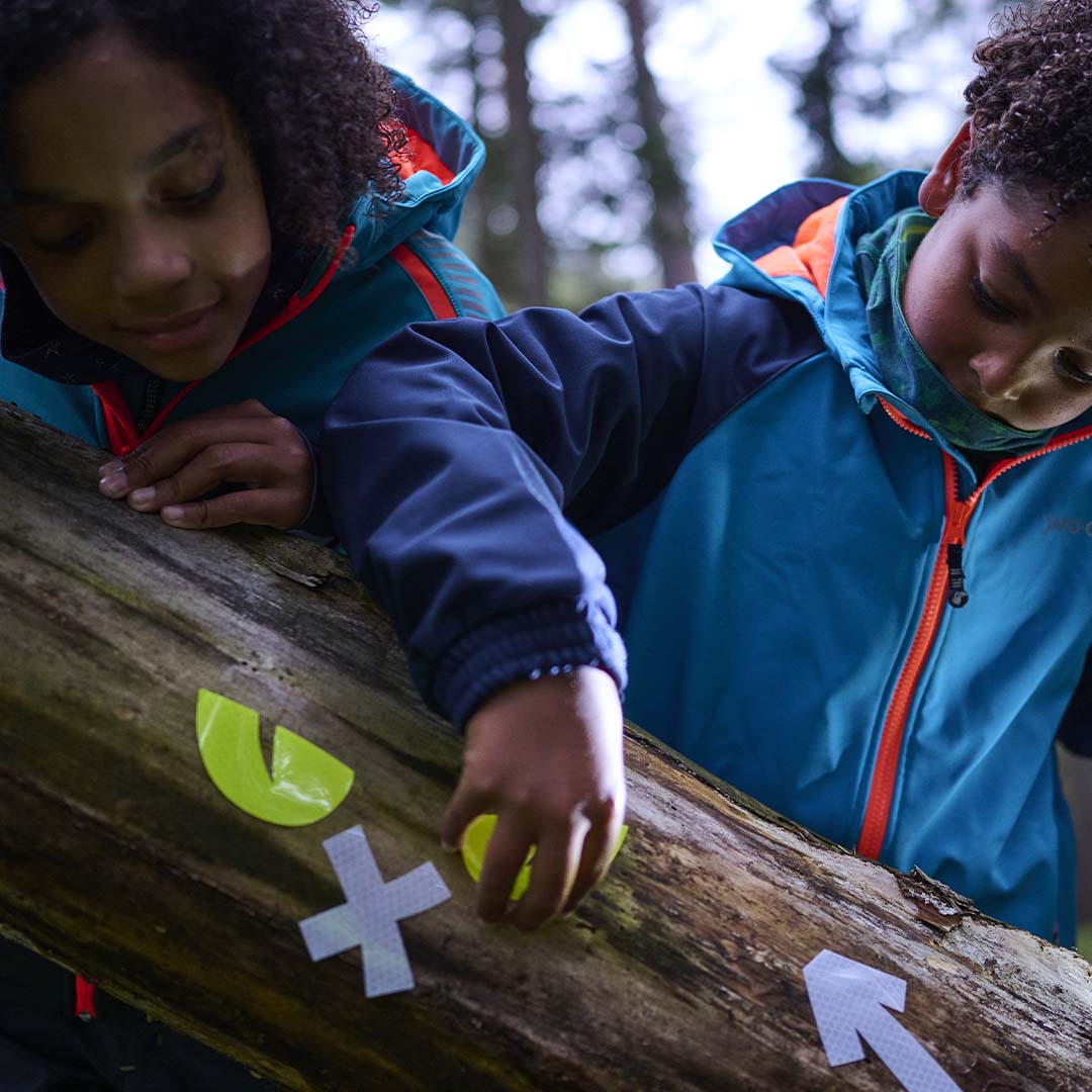 Twee kinderen plakken reflecterende stickers, waaronder ogen en een pijl, op een boomstam in het bos.