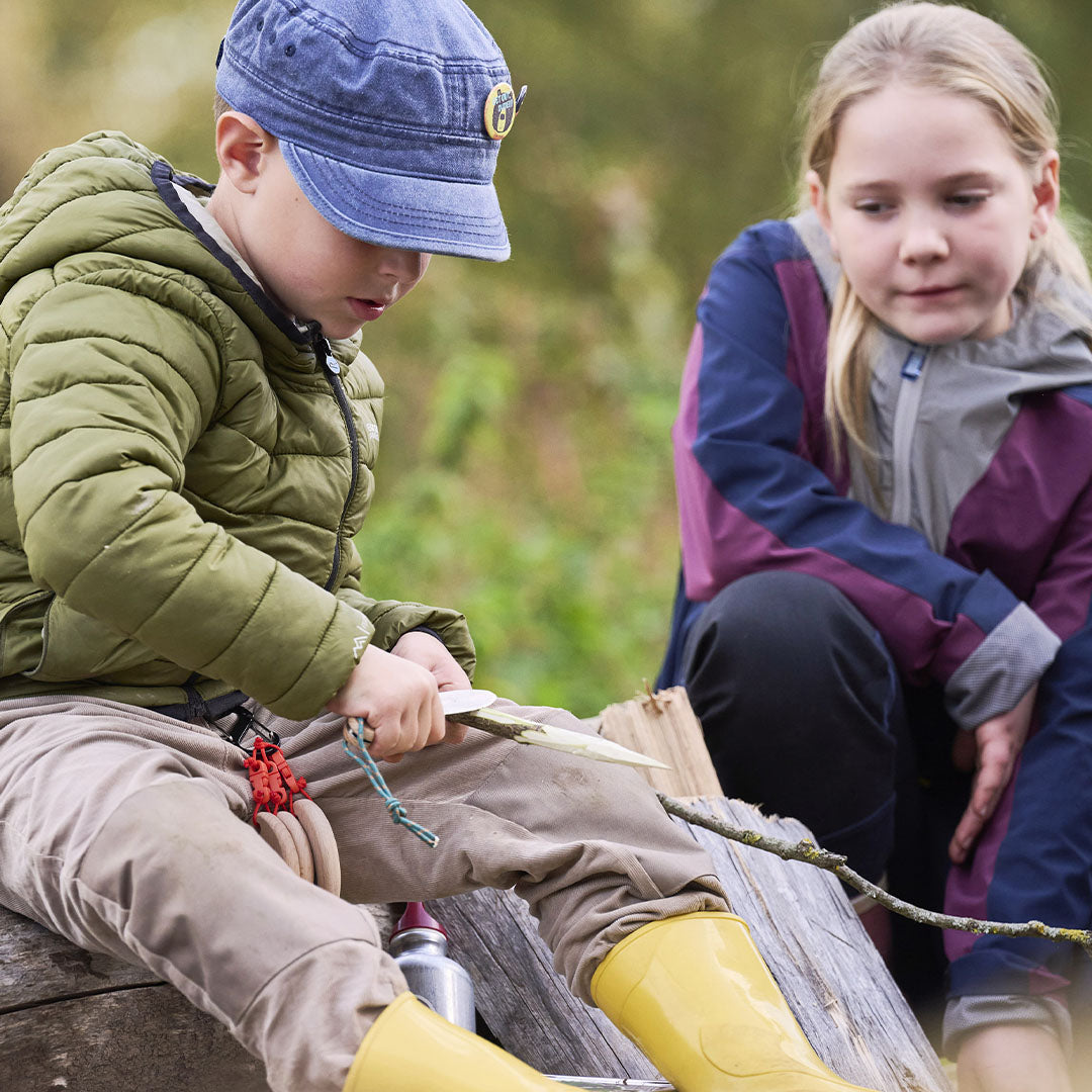 Een kind snijdt in een tak met het Haba Terra Kids zakmes, zittend op een boomstam.