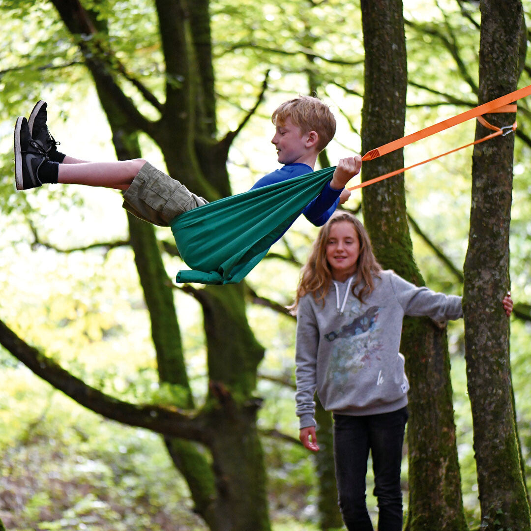 Een jongen schommelt in de groene Hape 2-in-1 hangmat tussen bomen, met een meisje ernaast.