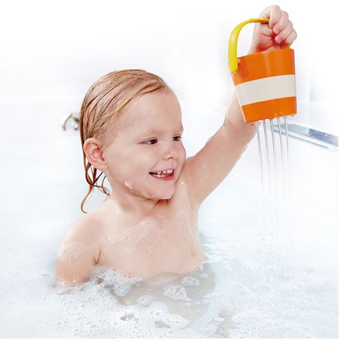 Een lachende peuter giet water uit een oranje Hape bademmertje met strepen in bad.