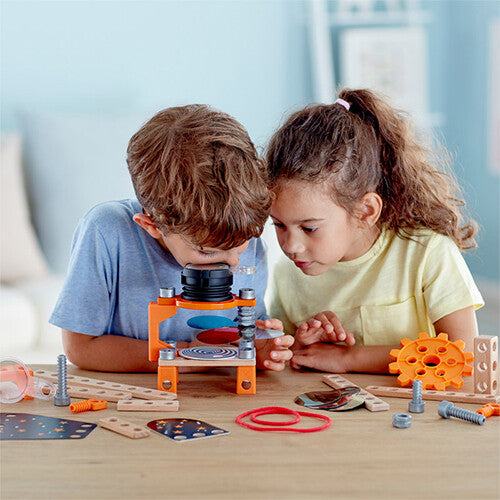 Twee kinderen kijken geconcentreerd naar een opstelling van de Hape bouwset optica laboratorium met gekleurde schijven.