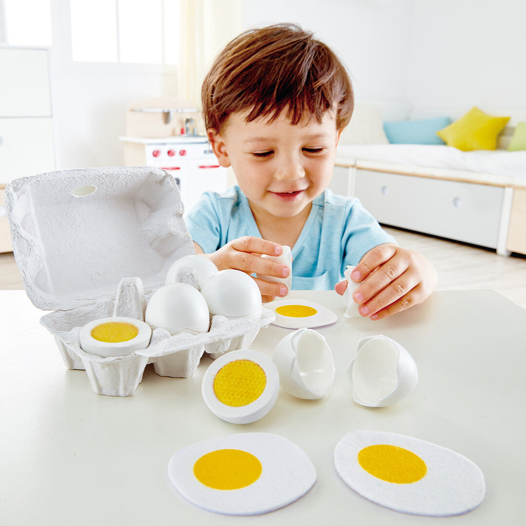 Een jongen speelt met de Hape speelgoed eieren, met een open eierdoos en losse eieren en eierschalen op tafel.