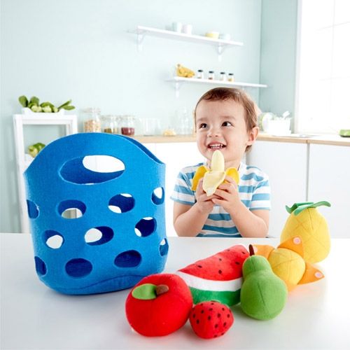 Een kind zit aan tafel met het Hape vilten fruitmandje en de fruitstukken, en houdt een gepelde banaan vast.