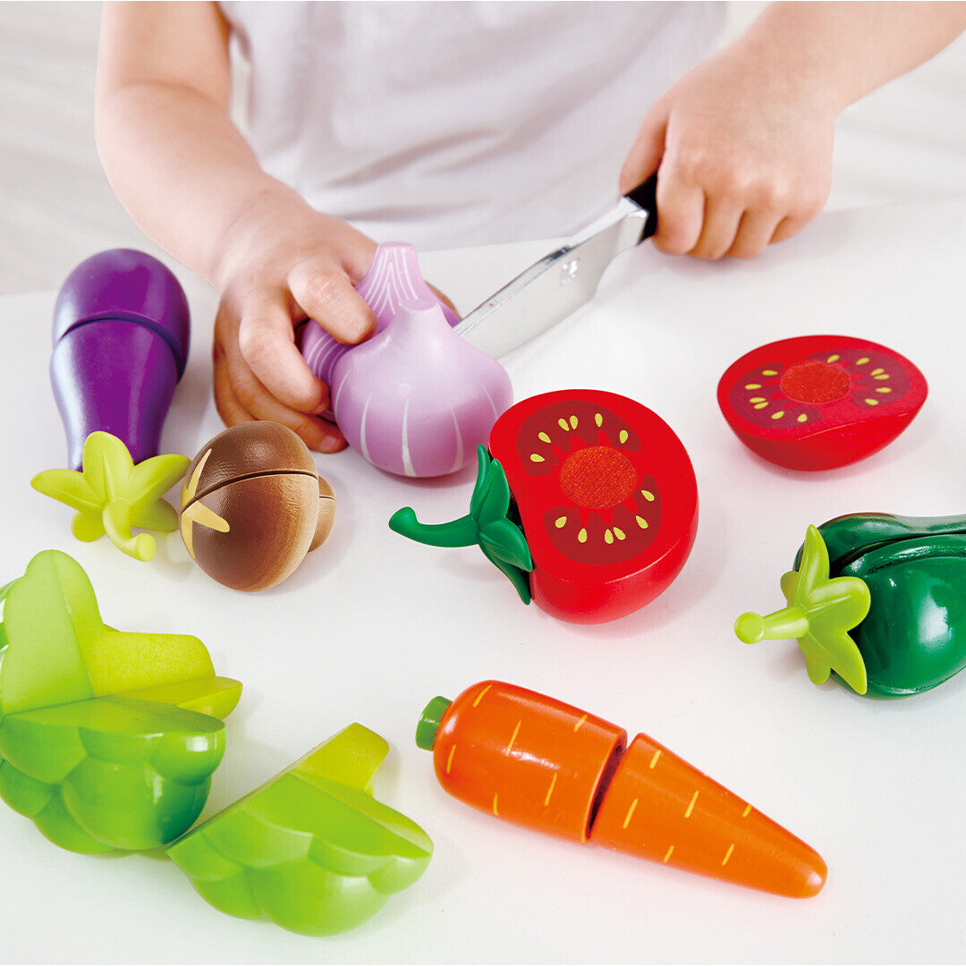 Kinderhanden snijden een Hape houten speelgoedknoflook met een speelgoedmes, omringd door andere speelgroenten.
