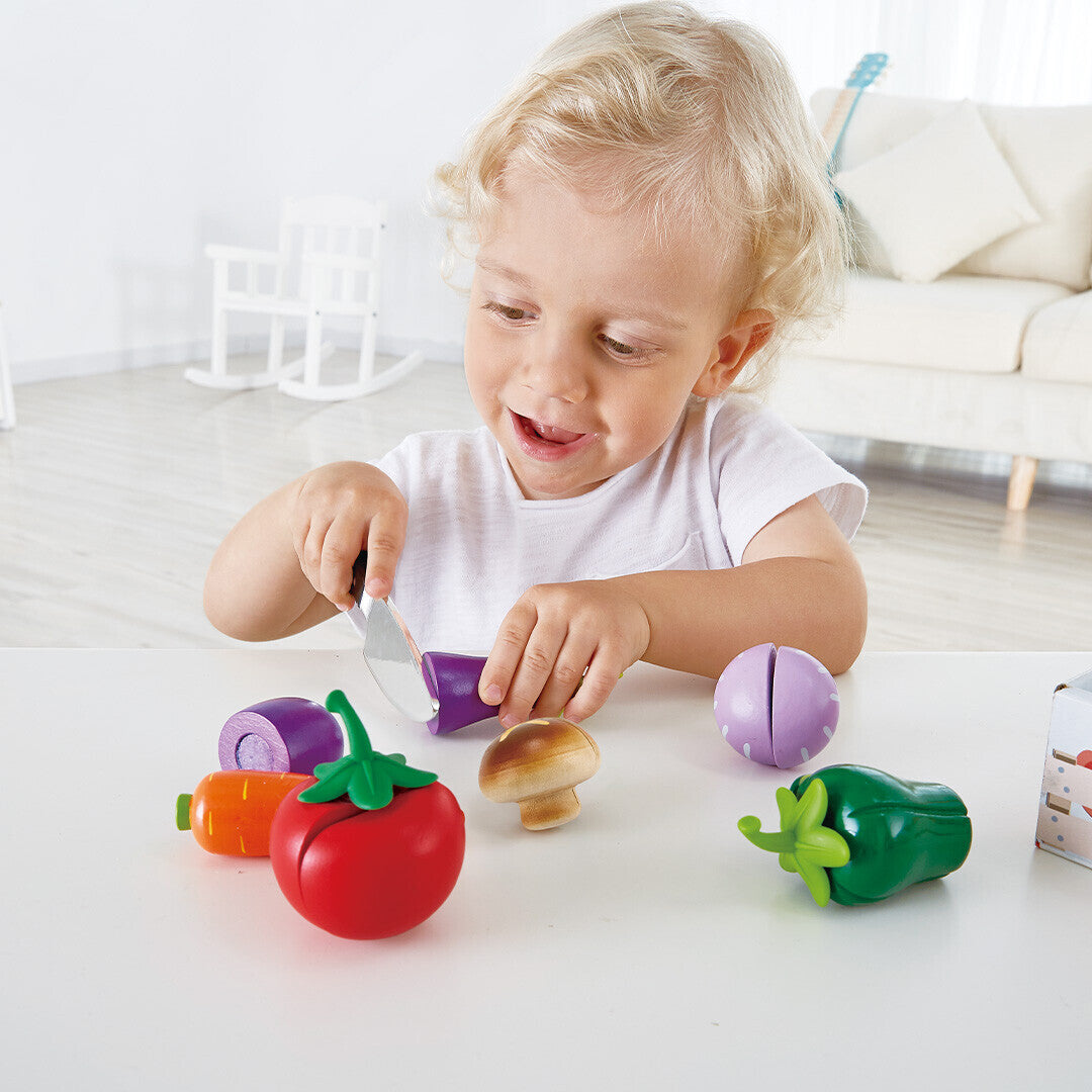 Een lachend kind snijdt een Hape houten speelgoedaubergine met een speelgoedmes, met andere speelgroenten op tafel.