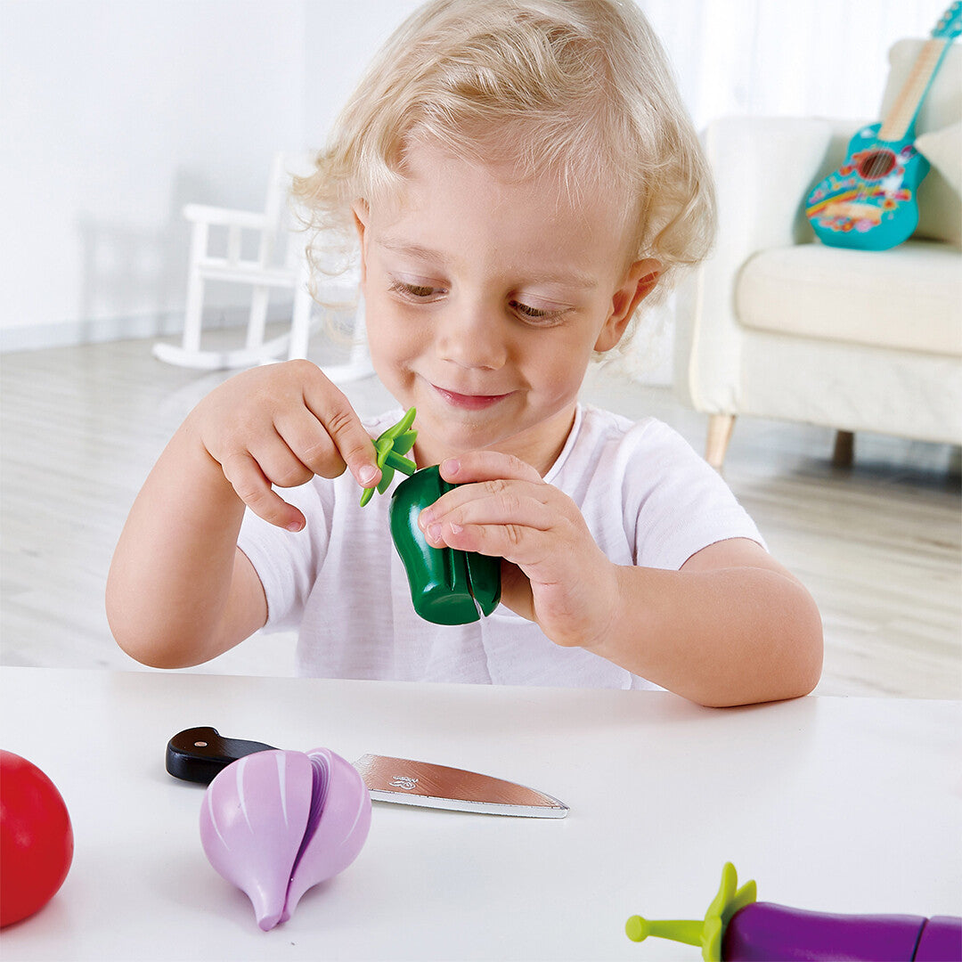 Een kind houdt een Hape houten speelgoedpaprika vast, met een speelgoedmes en andere speelgroenten op tafel.
