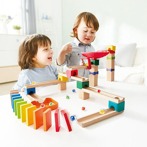 Twee lachende kinderen spelen met de Hape knikkerbaan en de kleurrijke dominostenen op een witte tafel.