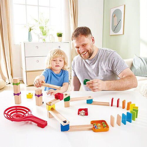 Een volwassen man en een kind spelen samen met de Hape knikkerbaan en de dominostenen op een witte tafel.