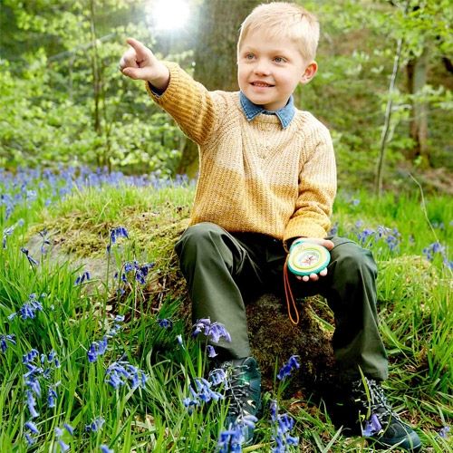 Een jongen zit in een bos met blauwe bloemen, houdt het Hape speelgoedkompas vast en wijst in de verte.