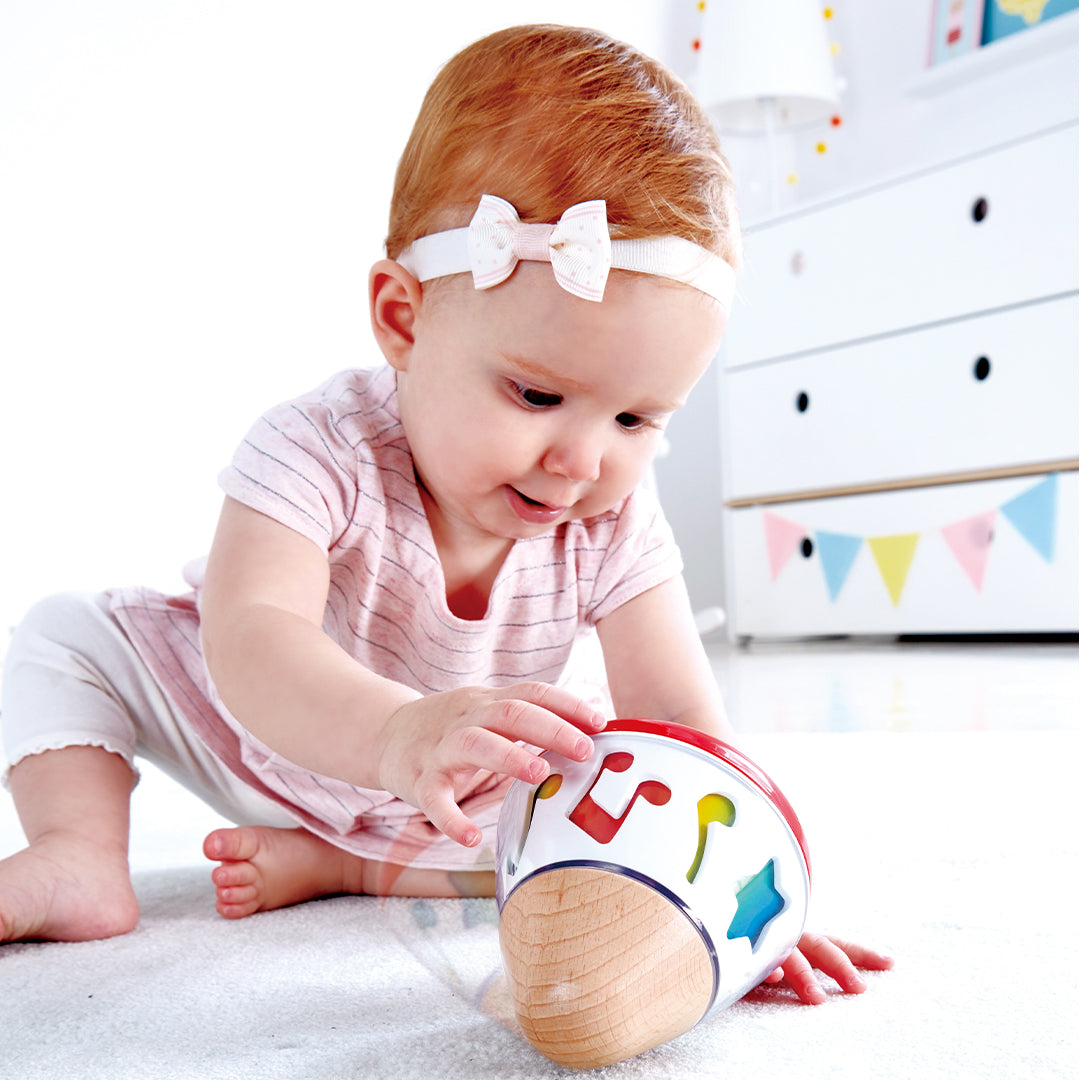 Een baby met een strik in het haar reikt naar het Hape muziekspeelgoed op een witte vloer.