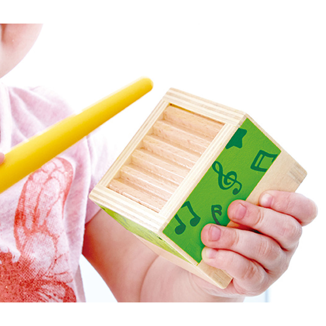 Detail van een kind dat een groen Hape muzikaal bouwblok met een wasbordstructuur vasthoudt, samen met een gele drumstick.