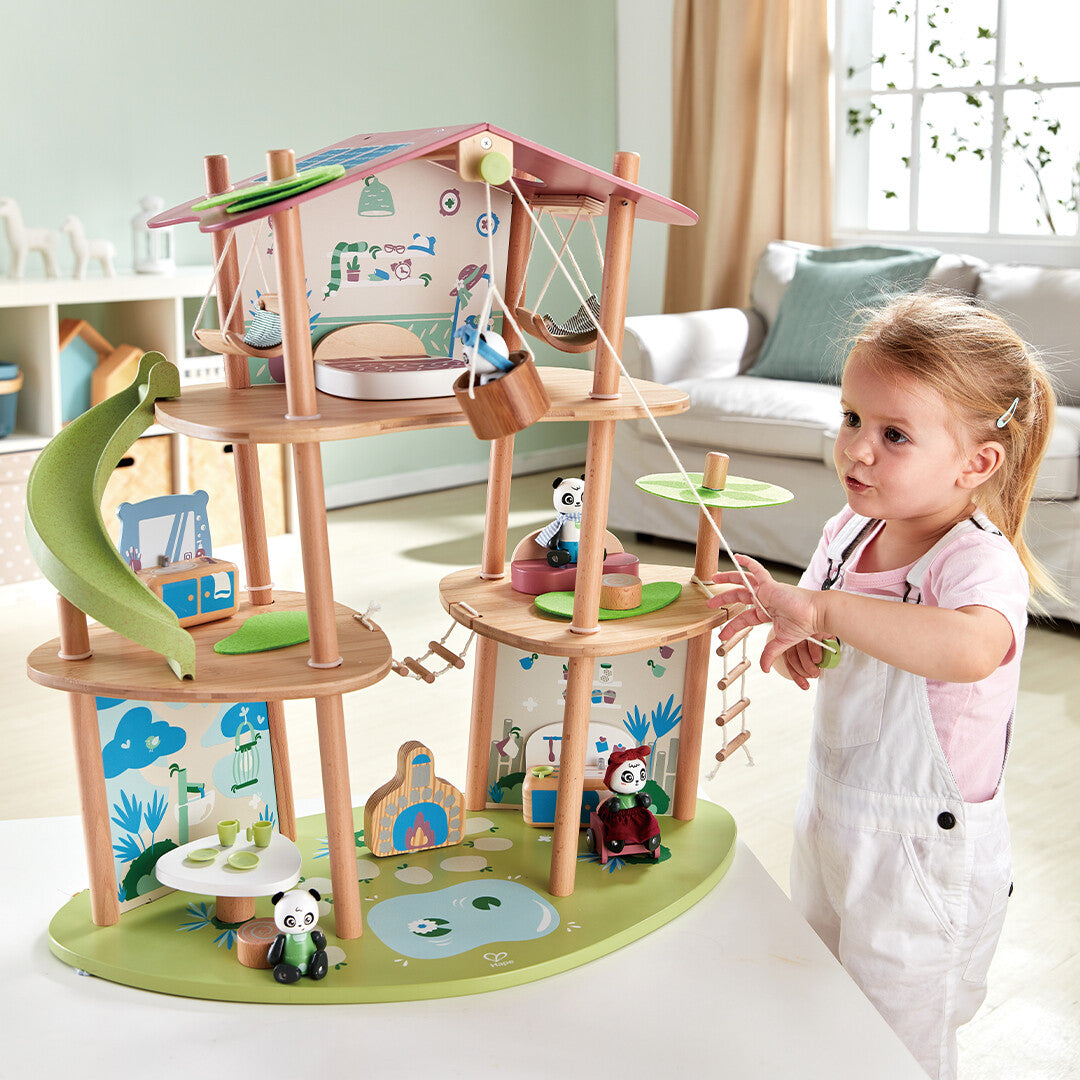 Een jong meisje speelt met het Hape houten poppenhuis in boomhutstijl met pandafamiliefiguren.