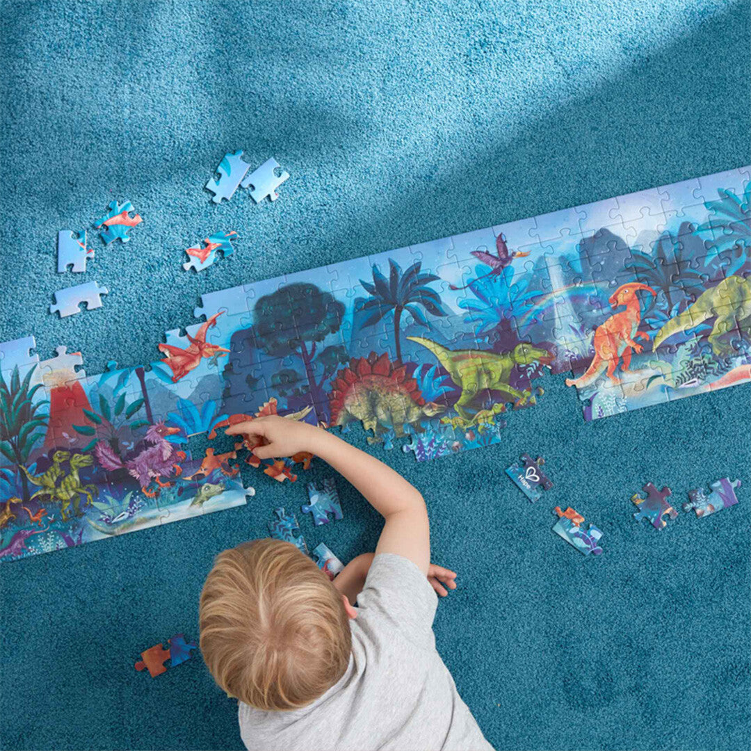 Een kind legt de Hape dinosaurus legpuzzel van 1,5 meter lang op een blauw tapijt.