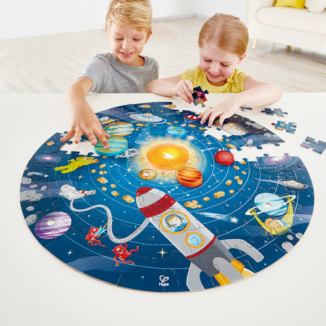 Twee kinderen leggen de Hape zonnestelsel legpuzzel met raket en planeten op een witte tafel.