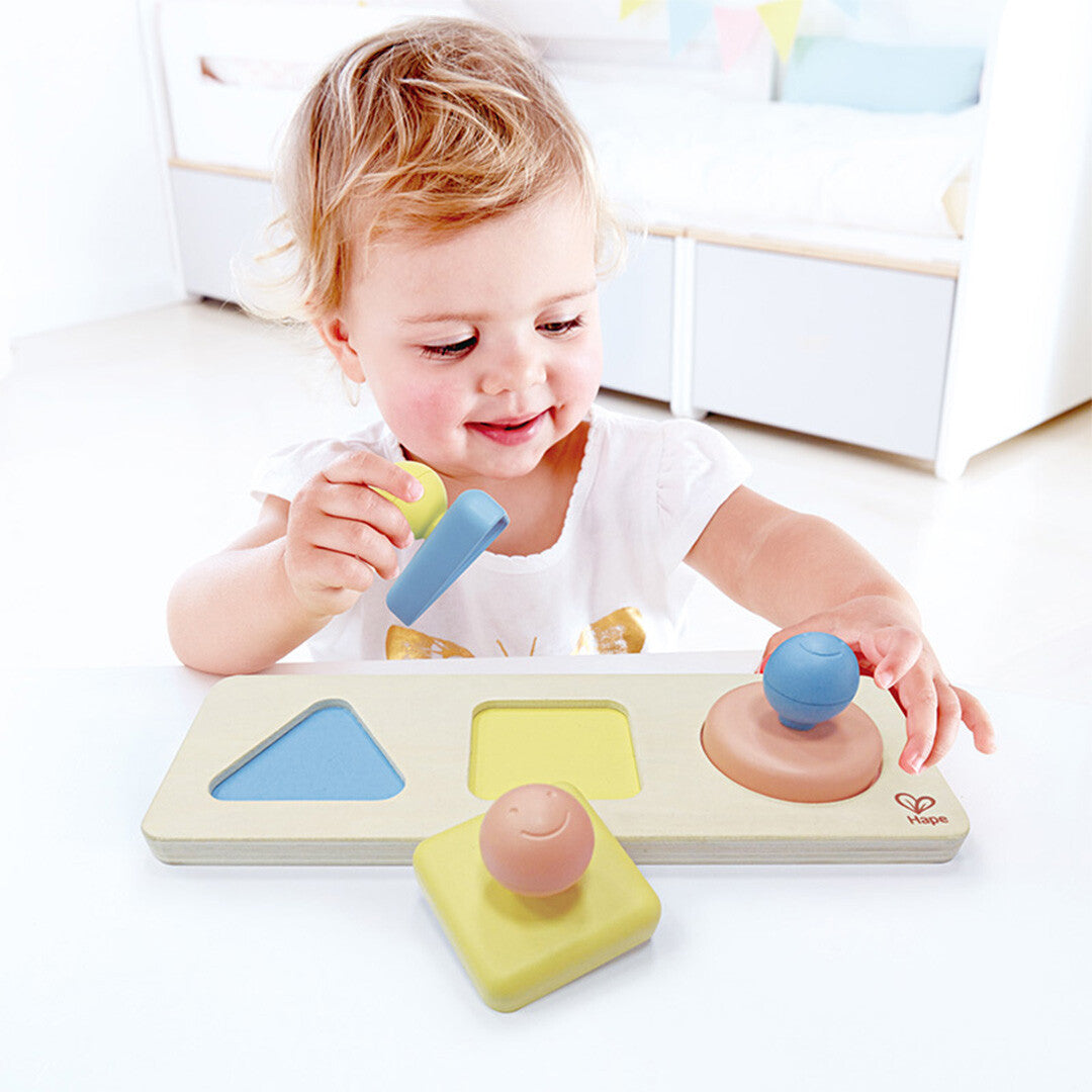 Een lachende peuter speelt met de Hape houten vormenpuzzel, waarbij een blauw puzzelstuk wordt vastgehouden.