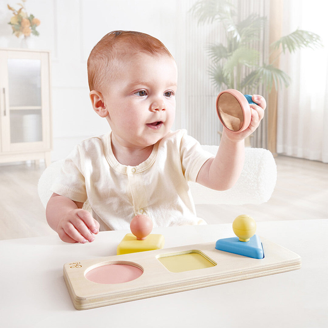 Een baby houdt een roze puzzelstuk met spiegel van de Hape houten vormenpuzzel omhoog.