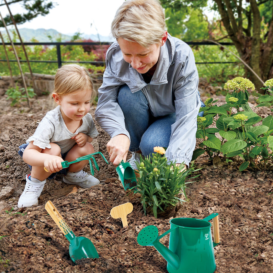 Een vrouw en een kind tuinieren samen met groene Hape tuingereedschappen in de aarde.