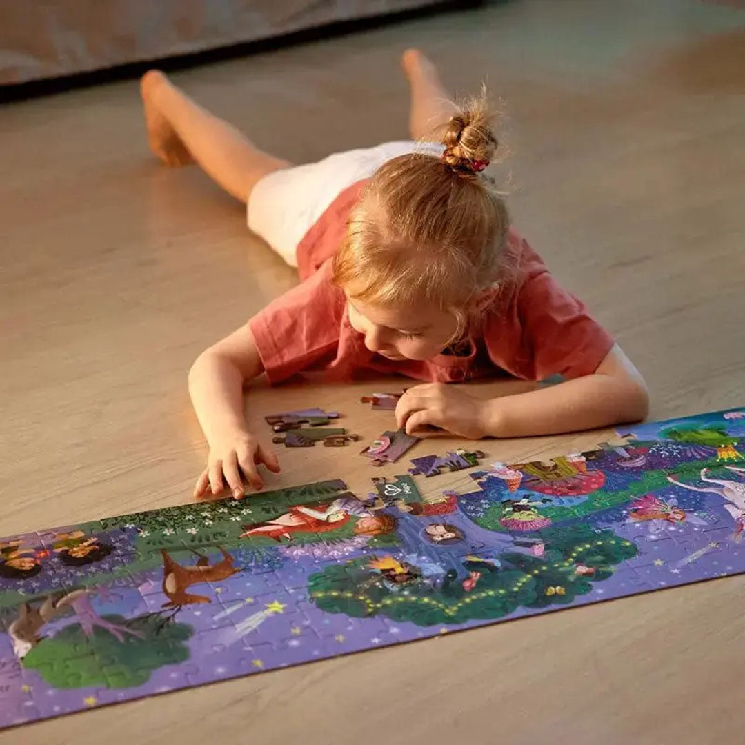 Een jong meisje ligt op de vloer en legt de Hape 'Magic Forest' vloerpuzzel met een magisch bos thema.