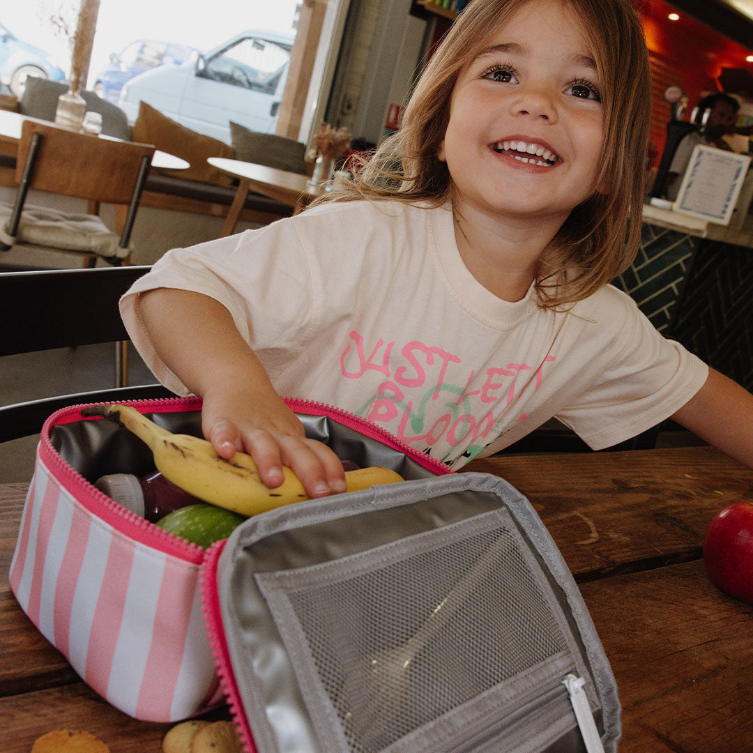 Een lachend kind zit aan tafel met de open hello hossy lunchtas met roze en witte strepen, gevuld met fruit.