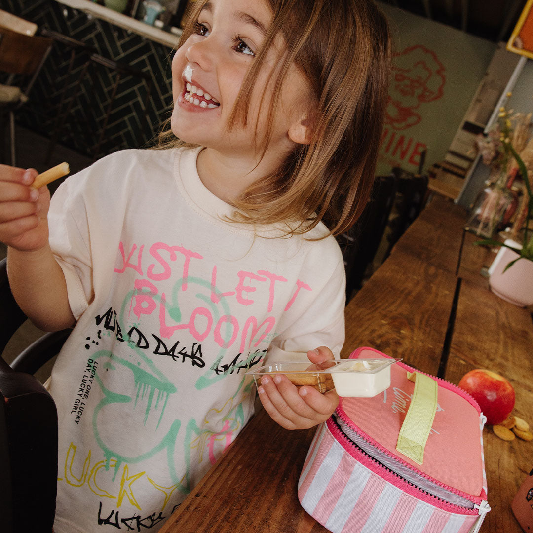 Een lachend kind eet snacks aan tafel, met de hello hossy lunchtas met roze en witte strepen ernaast.