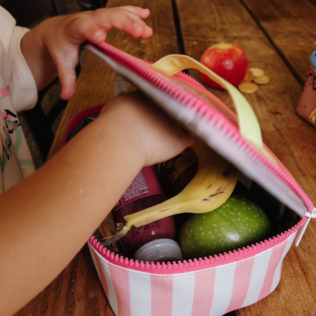 Een kind opent de hello hossy lunchtas met roze en witte strepen, gevuld met een banaan, appel en drinkfles.
