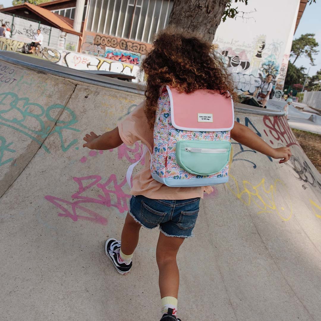 Een kind draagt de Hello Hossy veelkleurige schoolrugzak met happiness patroon op de rug in een skatepark.
