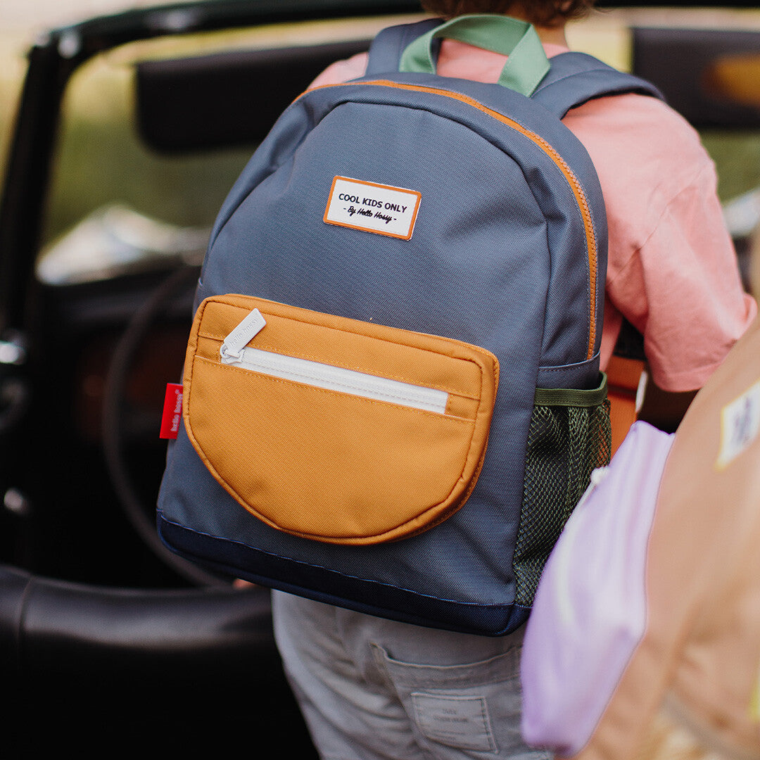 Een kind draagt de Hello Hossy schoolrugzak in marineblauw met oranje voorvak op de rug, met een auto op de achtergrond.