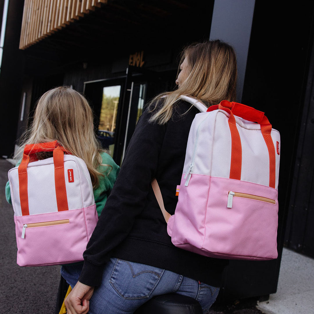 Twee personen, een volwassene en een kind, dragen elk een Hello Hossy schoolrugzak in roze en lichtroze op hun rug.