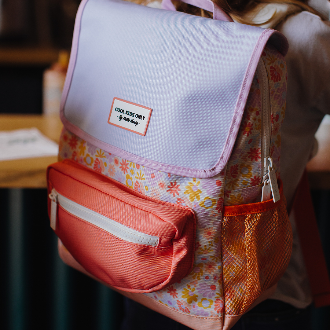 Kind draagt de Hello Hossy schoolrugzak met retro bloemenpatroon en lichtpaarse flap, schuin van achteren gezien.